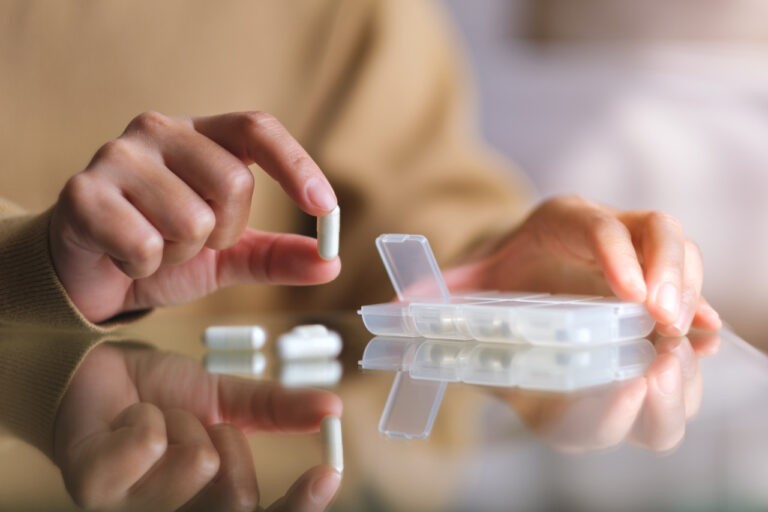 mãos de mulher segurando uma pílula em lado de uma caixinha de medicamentos em cima da mesa
