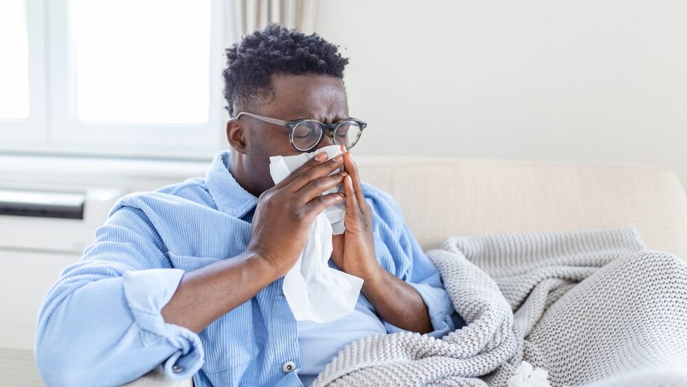 Homem sentado no sofá com um cobertor e assoando o nariz