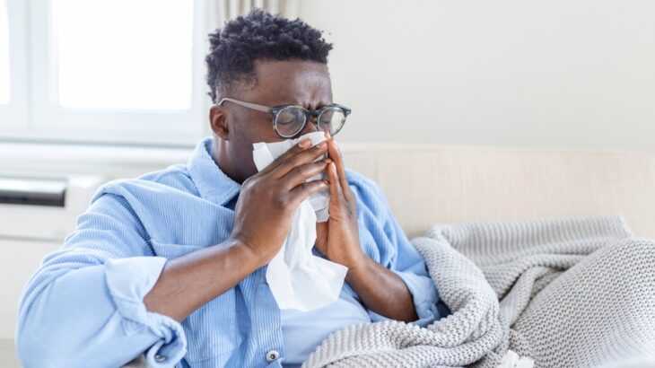 Homem sentado no sofá com um cobertor e assoando o nariz