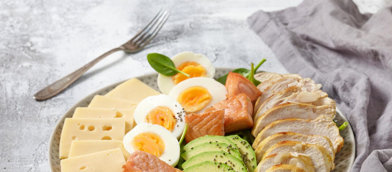 foto mostra um prato típico da dieta Atkins, com frango, ovos cozidos, abacate e salmão. Ao lado, um guardanapo de pano e um garfo