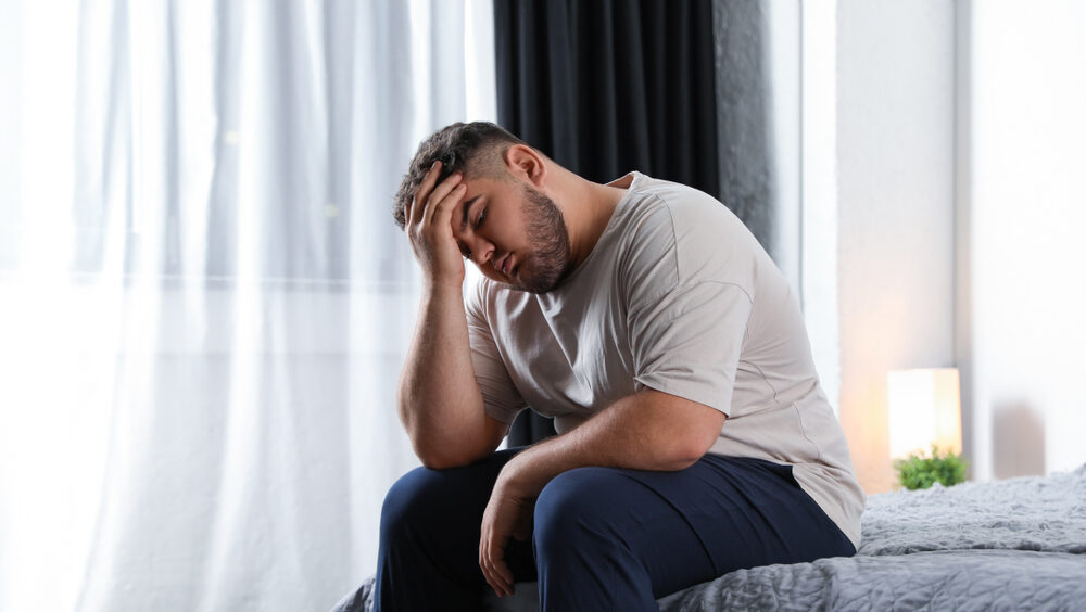 Homem com sobrepeso sentado na cama e com semblante preocupado