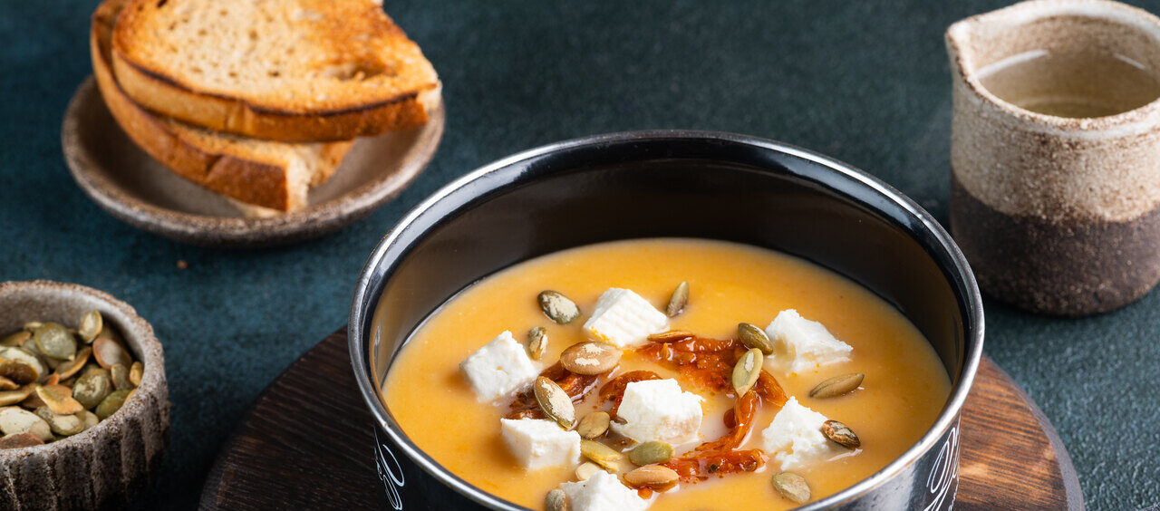 foto de uma cumbuca com sopa de abóbora, sementes de abóbora e pedaços de queijo. Ao lado encontram-se fatias de pão, mais sementes de abóbora, azeite e uma colher