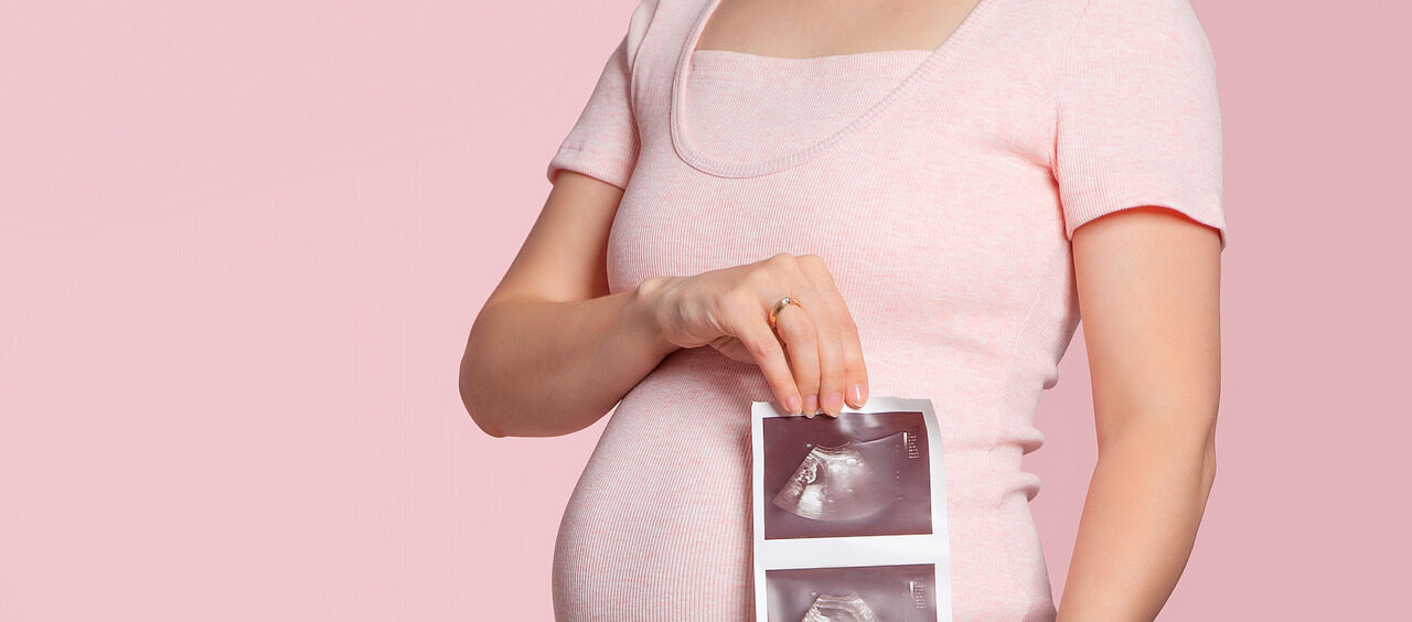 foto de uma grávida com 22 semanas de gestação da barriga para baixo. Ela segura fotos do ultrassom do bebê