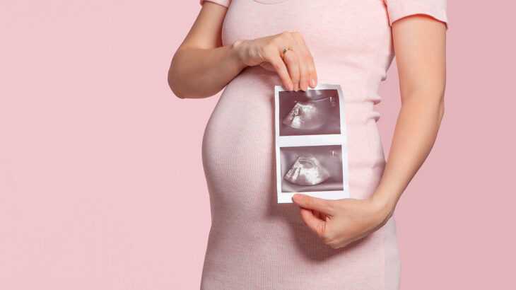 foto de uma grávida com 22 semanas de gestação da barriga para baixo. Ela segura fotos do ultrassom do bebê