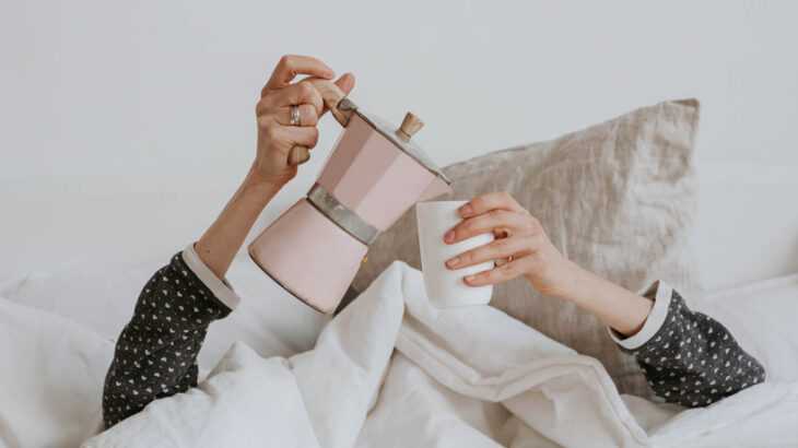pessoa deitada na cama com a cabeça coberta e as mãos aparentes, segurando uma cafeteira e uma xícara