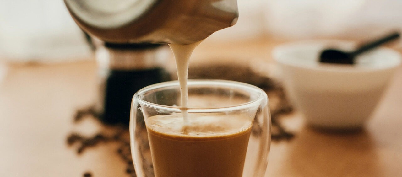 foto de um copo de capuccino sendo servido em cima de uma mesa com grãos de café e um açucareiro ao lado
