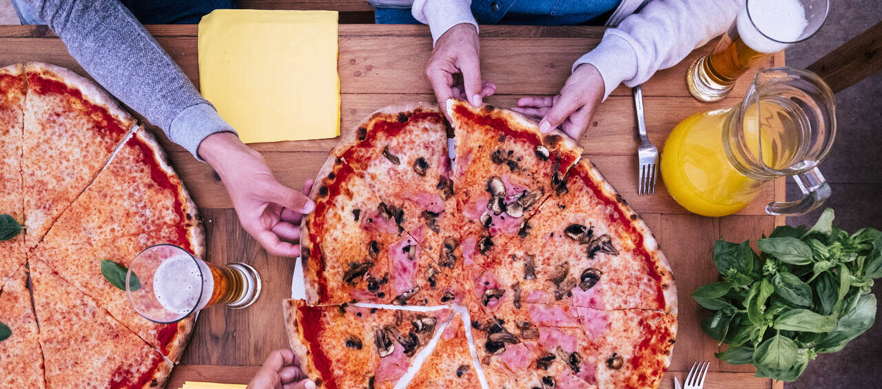 mesa vista de cima com uma pizza bem grande e mãos pegando as fatias