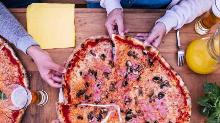 mesa vista de cima com uma pizza bem grande e mãos pegando as fatias