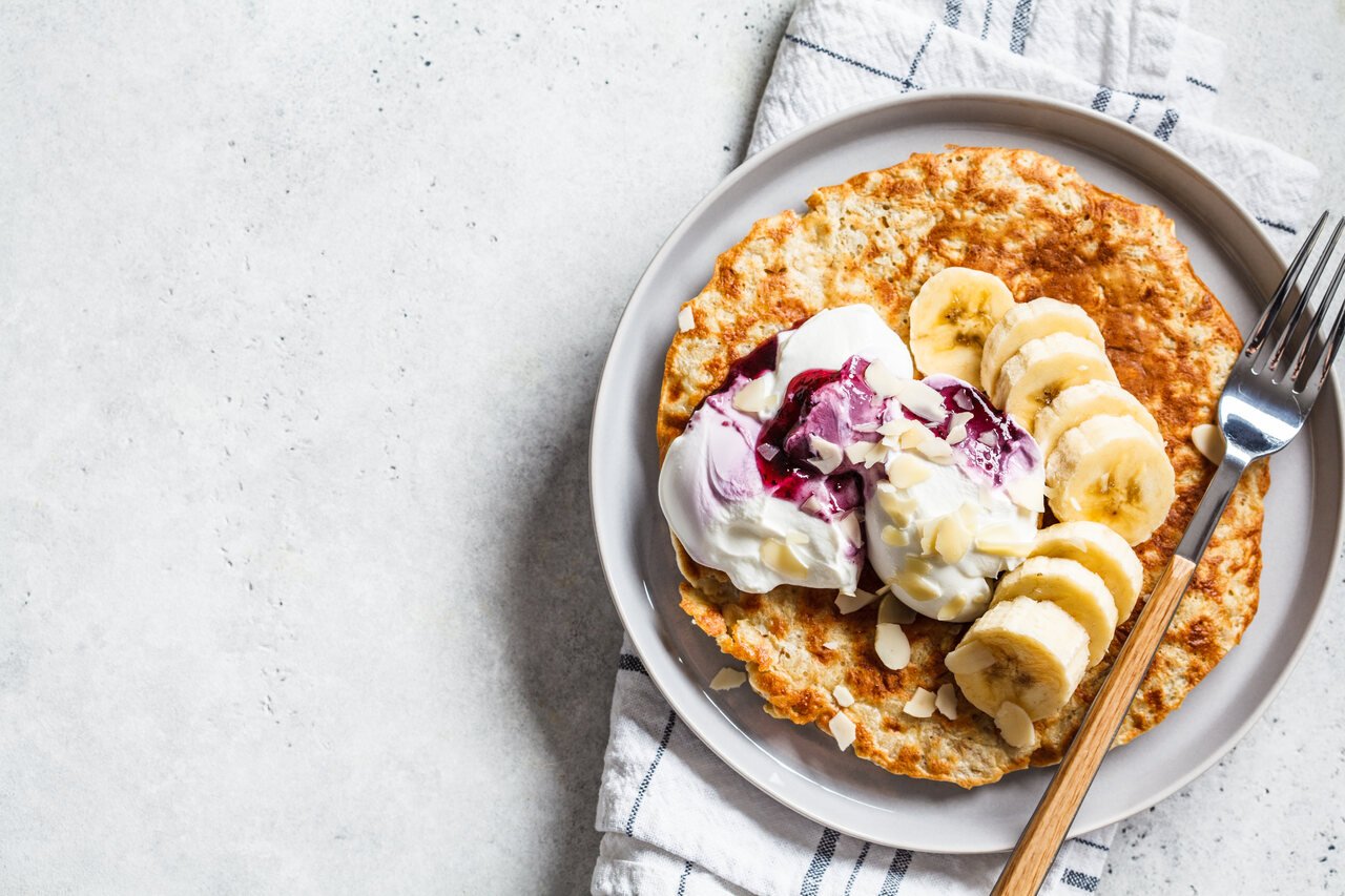 foto de um prato com uma panqueca de banana e aveia, em cima um creme de iogurte e mirtilo e umas bananas fatiadas