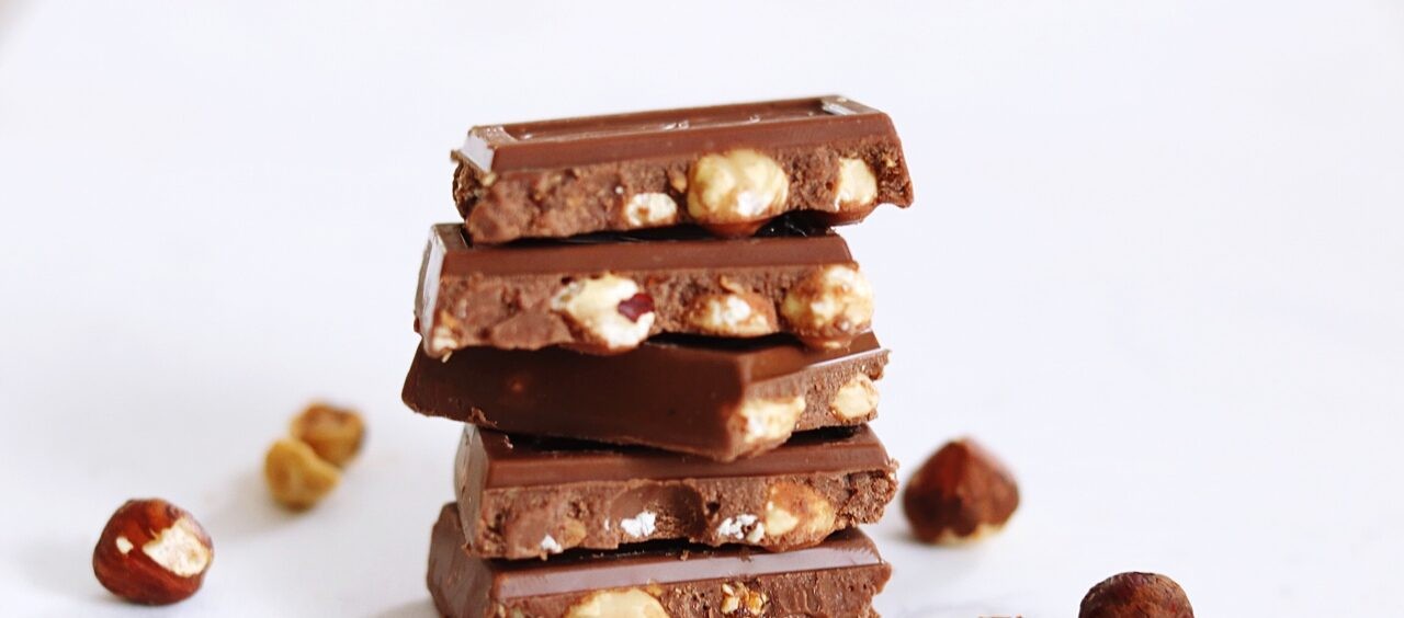 foto de quadradinhos de chocolate com pedaços de avelã, um em cima do outro. Ao lado, avelãs jogadas