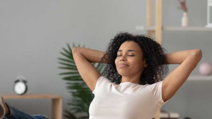 mulher negra sentada na sala de olhos fechados e relaxando com os braços atrás da cabeça