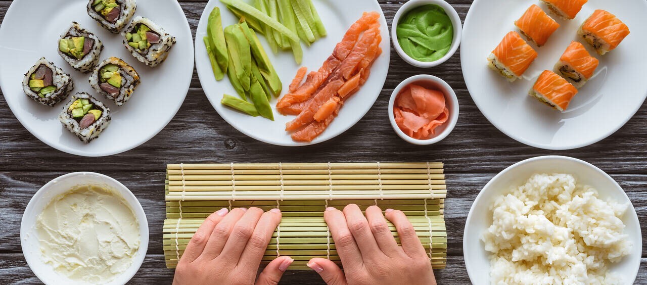 foto vista de cima de mãos enrolando um rolo de sushi na esteira de bambu, e ao lado peças de sushi, arroz japonês e outros ingredientes do prato