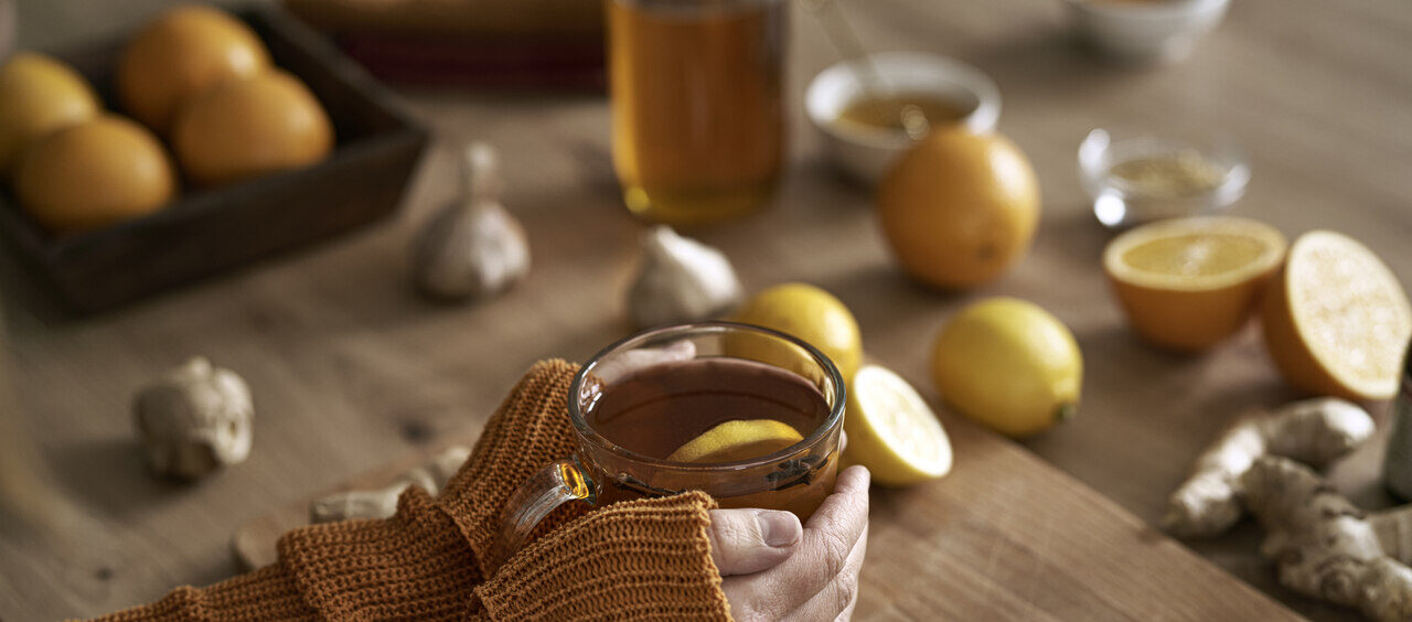 mãos segurando uma xícara de chá em cima de uma tábua de madeira com gengibre, alho, limões cortados e mel ao lado