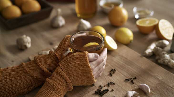 mãos segurando uma xícara de chá em cima de uma tábua de madeira com gengibre, alho, limões cortados e mel ao lado