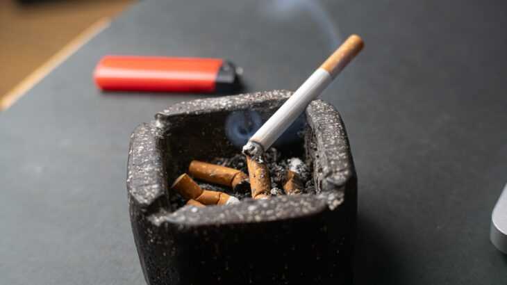 foto de um cinzeiro cheio de bitucas, um cigarro aceso e um isqueiro ao lado