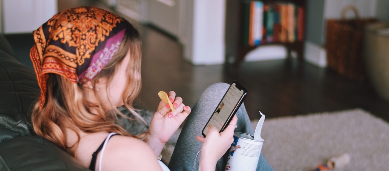 mulher de costas sentada no sofá mexendo no celular e comendo salgadinhos