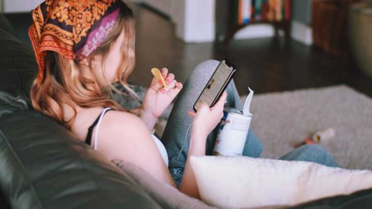 mulher de costas sentada no sofá mexendo no celular e comendo salgadinhos