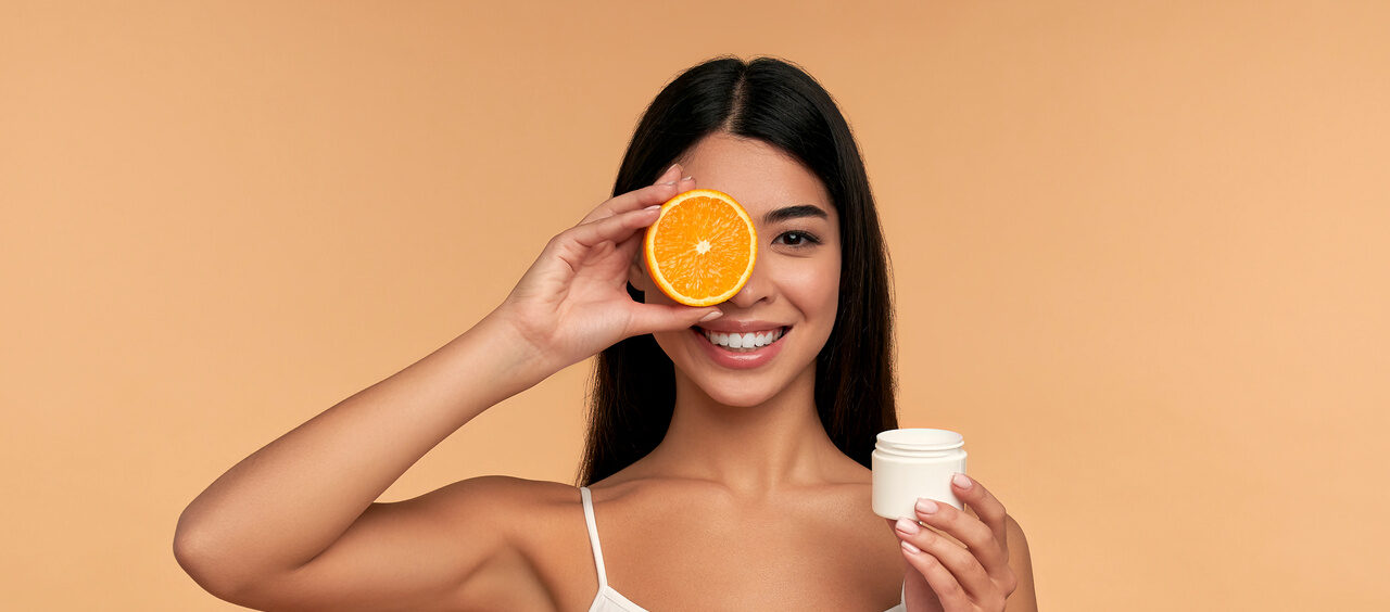mulher sorridente segurando uma laranja cortada ao meio em uma mão e um pote de creme facial em outra