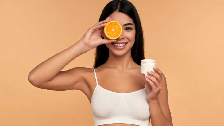 mulher sorridente segurando uma laranja cortada ao meio em uma mão e um pote de creme facial em outra