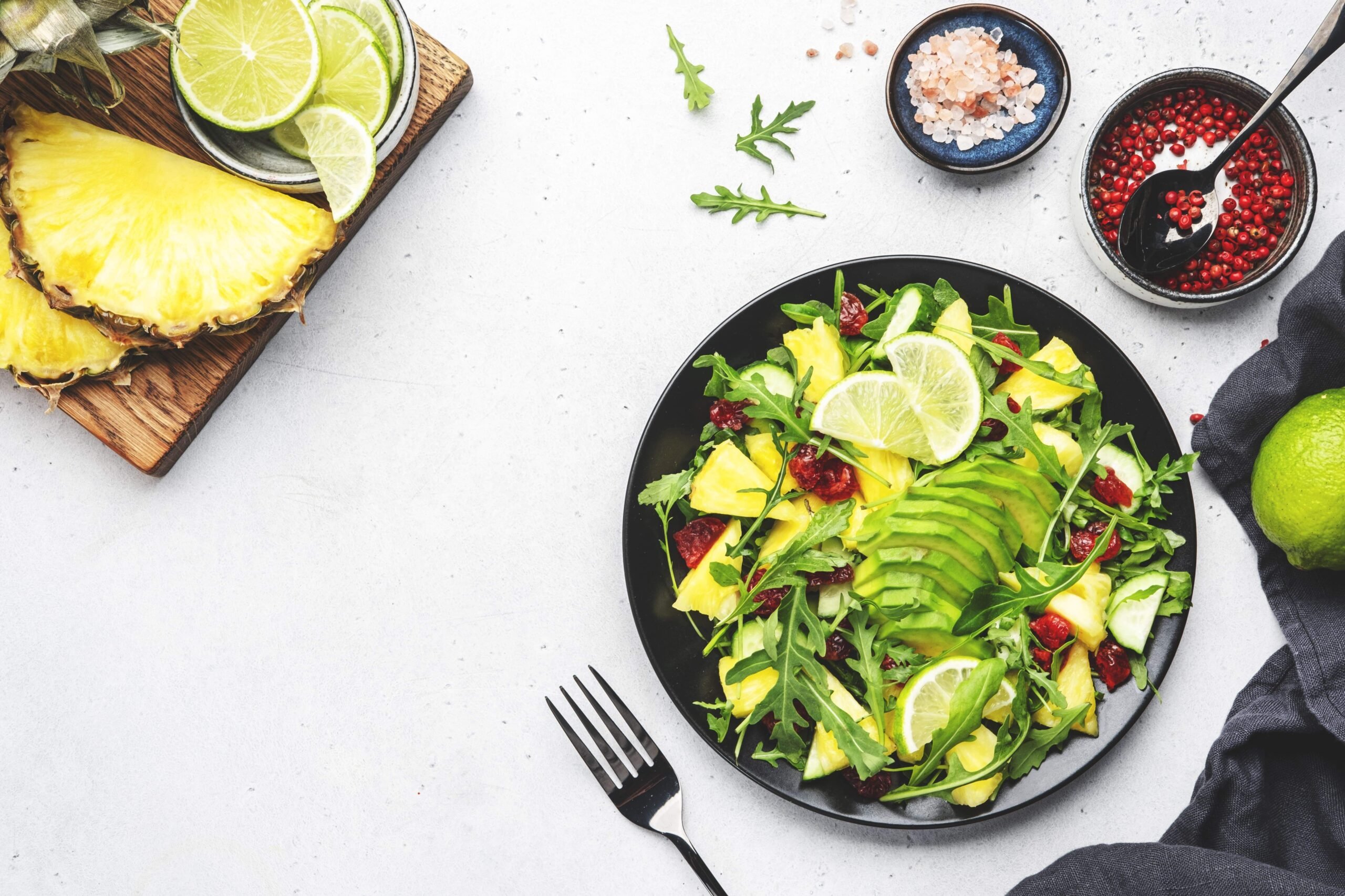 foto de um prato com salada de folhas e abacaxi visto de cima. Ao lado, molhos, garfo, um abacaxi cortada ao meio