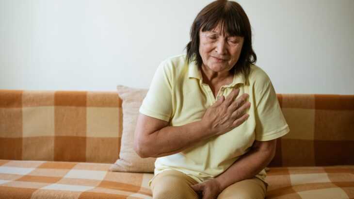 mulher idosa sentada no sofá com a mão no peito e o semblante de dor