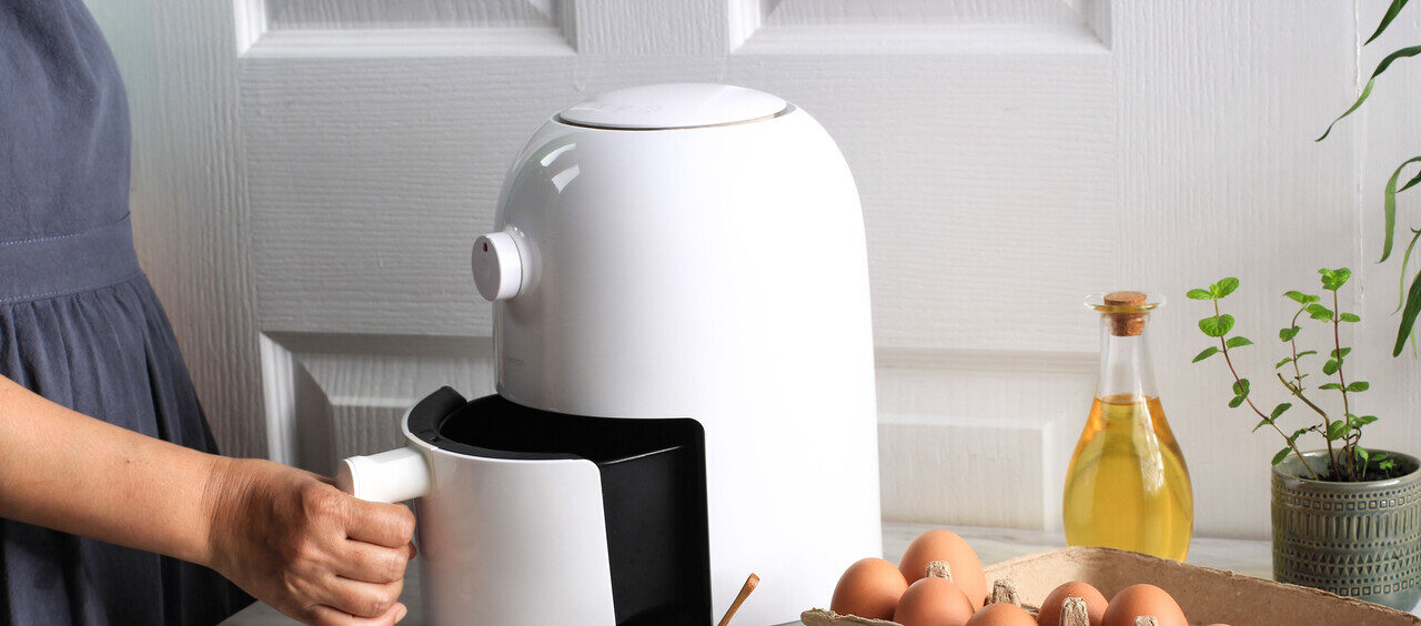 foto mostra uma mão abrindo uma air fryer branca, ao lado encontram-se ingredientes como ovos e bananas descascadas