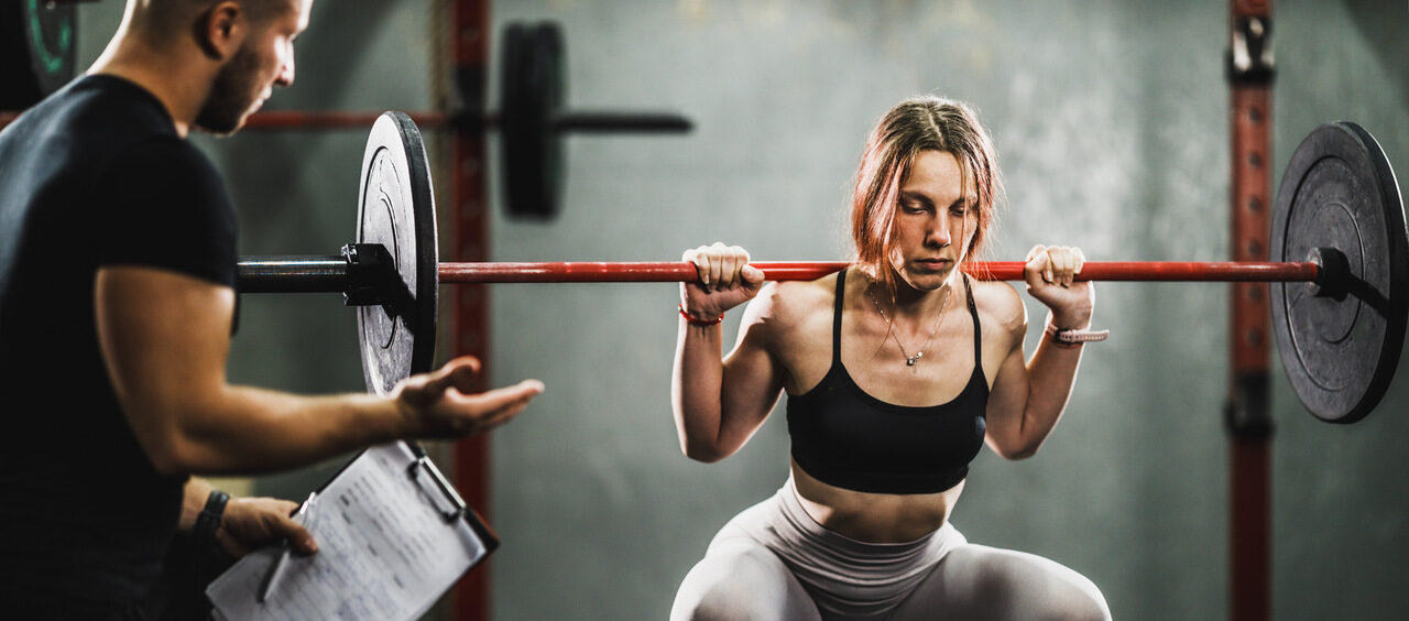 mulher fazendo um agachamento na academia ao lado do personal