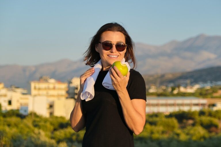 mulher madura comendo uma maçã ao ar livre. Ela está de óculos de sol e tem uma toalha apoiada nos ombros