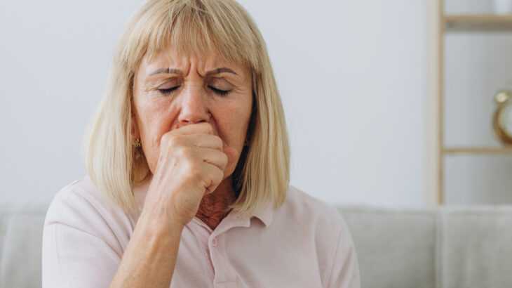 mulher idosa sentada no sofá da sala tossindo com a mão sobre a boca