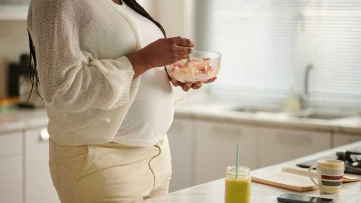 mulher grávida comendo um pote de iogurte com frutas na cozinha