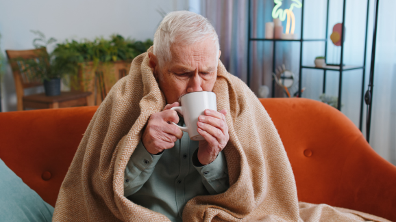 homem idoso sentando no sofá, com uma coberta sobre os ombros, tomando chá verde - um dos alimentos que aquecem o corpo