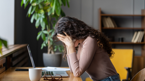 mulher sentada na frente de mesa e computador, apoiando os cotovelos na mesa e com a cabeça entre as mãos. produtividade e saúde mental estão conectadas.